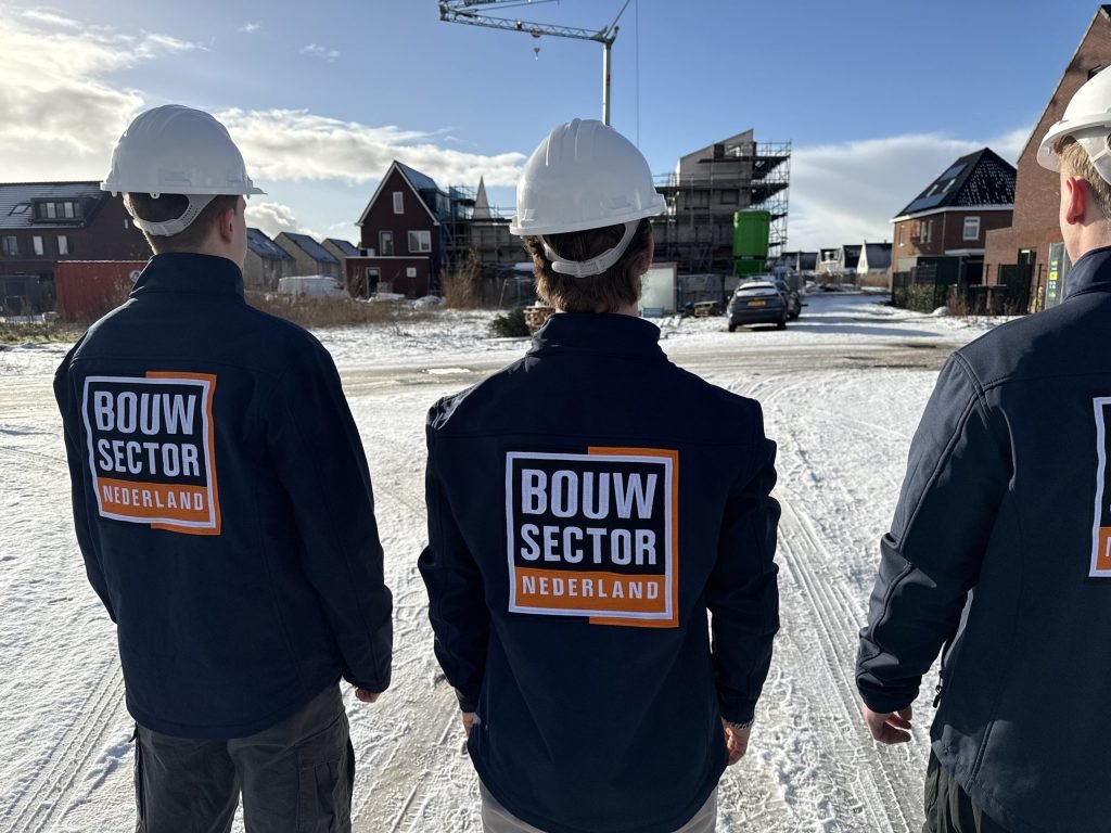 Drie vakmensen van Bouwsector Nederland met witte helm in een besneeuwde straat met huizen in aanbouw